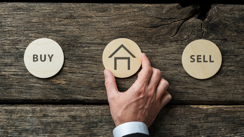 Real estate market conceptual image - hand of a businessman placing wooden cut circle with house shape on it in between Buy and Sell signs.